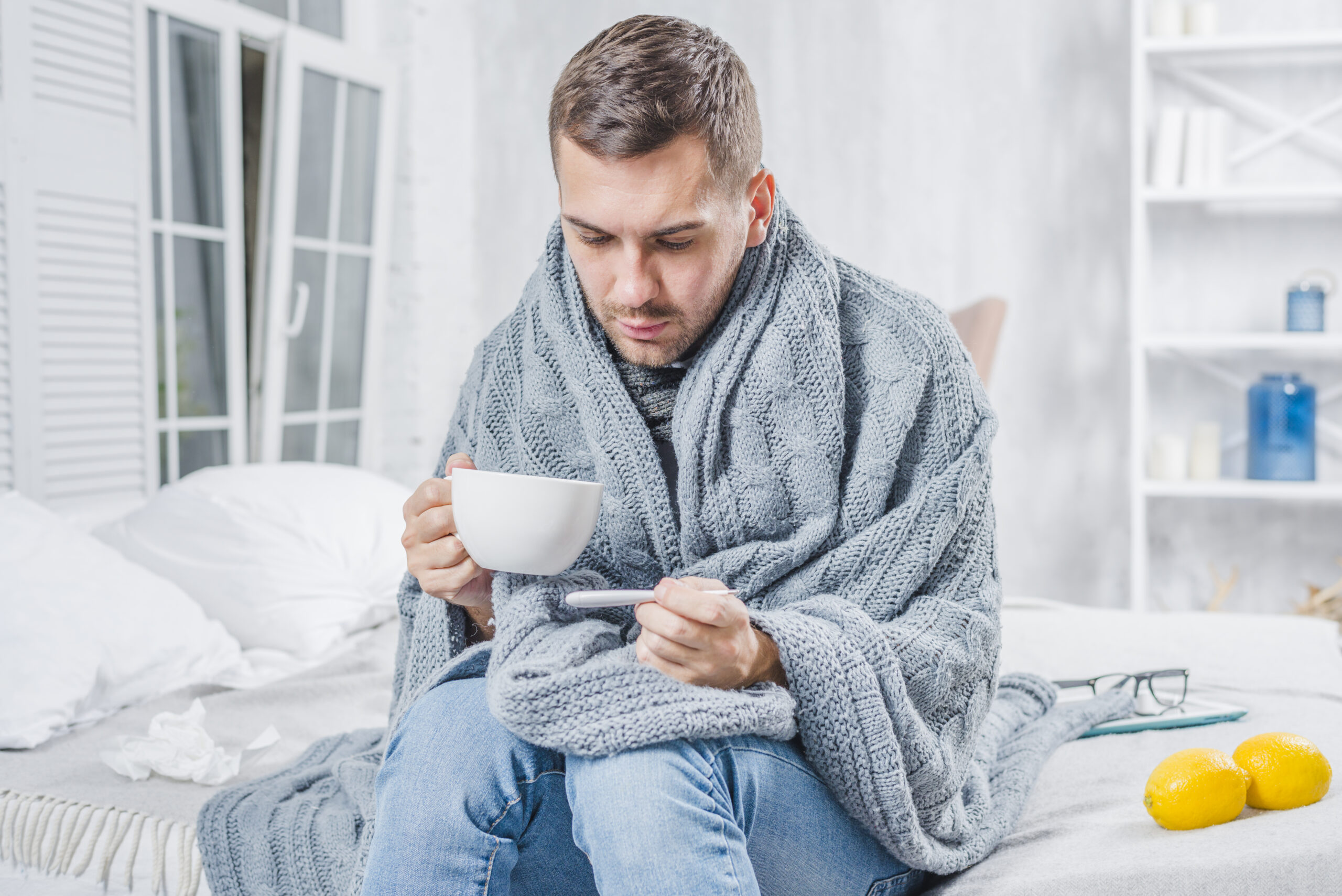 Bolesni muškarac sjedi u krevetu, drži šalicu čaja i provjerava temperaturu toplomjerom.